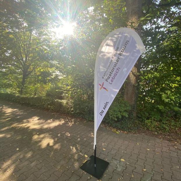 Eine schmale gepflasterte Straße führt durch einen grünen, schattigen Bereich mit vielen Bäumen und Sträuchern. Rechts im Vordergrund steht eine hohe, weiße, tropfenförmige Werbefahne auf einem schwarzen Fuß. Auf der Fahne steht gut sichtbar der Text „Katholische Kirche Pastoraler Raum Lebach“ sowie der Slogan „Da sein.“ und ein kleines Kreuzsymbol. Die Sonne scheint von links durch die Baumkronen und erzeugt helle Lichtstrahlen und starke Lichtreflexe. Der Boden ist teilweise im Schatten, teilweise von Sonnenlicht beleuchtet. Im Hintergrund sind dichte Büsche und weitere Bäume zu sehen, die eine ruhige, natürliche Umgebung bilden. (c) PastR Lebach Eine schmale gepflasterte Straße führt durch einen grünen, schattigen Bereich mit vielen Bäumen und Sträuchern. Rechts im Vordergrund steht eine hohe, weiße, tropfenförmige Werbefahne auf einem schwarzen Fuß. Auf der Fahne steht gut sichtbar der Text „Katholische Kirche Pastoraler Raum Lebach“ sowie der Slogan „Da sein.“ und ein kleines Kreuzsymbol. Die Sonne scheint von links durch die Baumkronen und erzeugt helle Lichtstrahlen und starke Lichtreflexe. Der Boden ist teilweise im Schatten, teilweise von Sonnenlicht beleuchtet. Im Hintergrund sind dichte Büsche und weitere Bäume zu sehen, die eine ruhige, natürliche Umgebung bilden.
