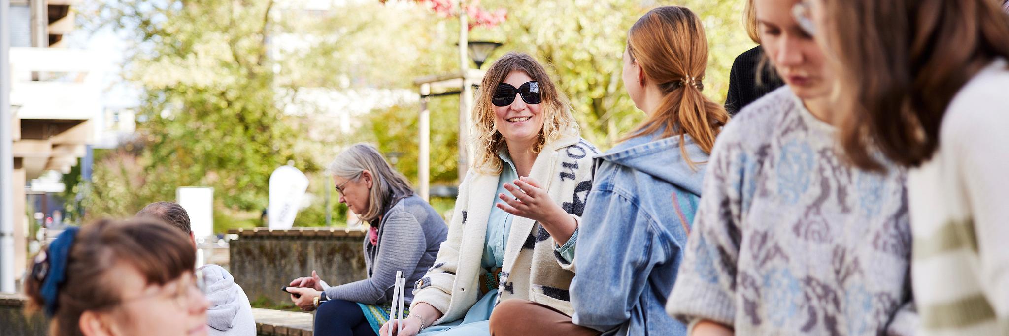Mehrere Frauen sitzen auf stufenförmig angeordneten Holzbänken. Im Vordergrund drei Personen, die sich unterhalten und lächeln. Im Hintergrund zwei Frauen, die sich unterhalten, eine davon trägt eine schwarze Brille und einen Langstock. (c) Anna Spindelndreier | helloyou.studio | Gesellschaftsbilder.de Mehrere Frauen sitzen auf stufenförmig angeordneten Holzbänken. Im Vordergrund drei Personen, die sich unterhalten und lächeln. Im Hintergrund zwei Frauen, die sich unterhalten, eine davon trägt eine schwarze Brille und einen Langstock.